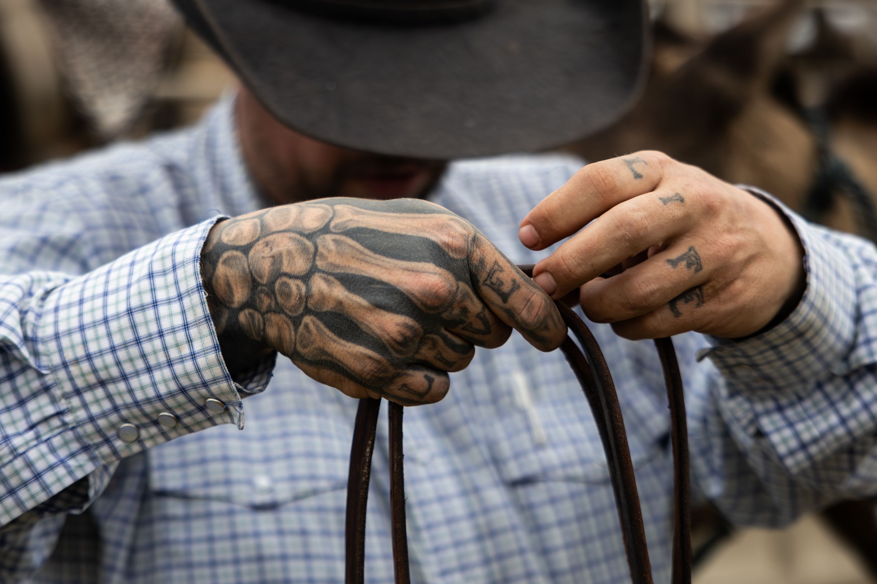 A close-up shot of a man's tattooed hands holding a rope. From left to right on the knuckles the words live and life are tattooed. His right hand is also heavily tattooed to look skeletal. The man is seen in the background from the torso up, though his face is hidden as he looks down and the brim of his cowboy hat hides it. This is Mason LaDue.