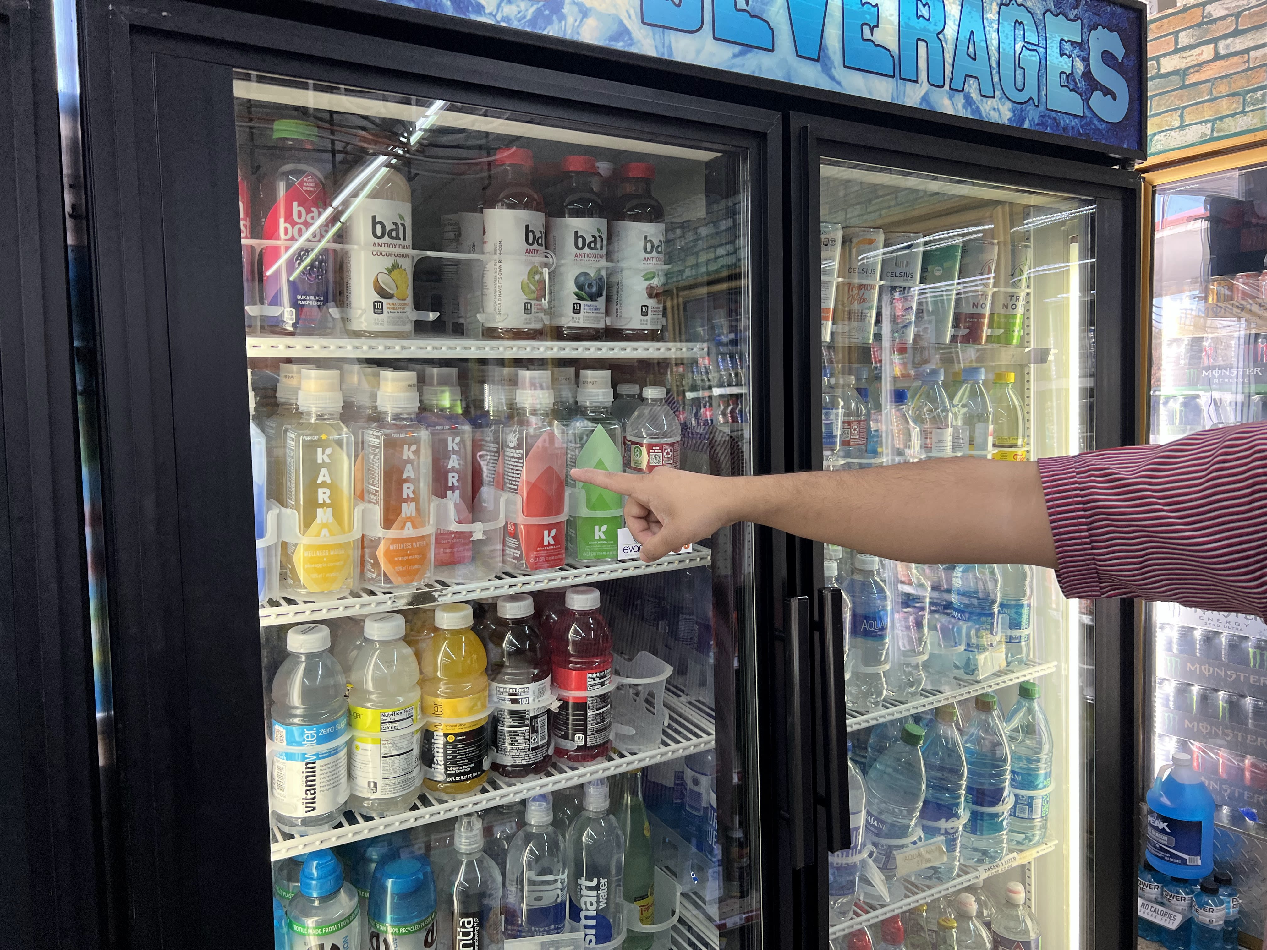 A hand is seen pointing to a refrigerated display case in a convenience store, from which can be seen several beverages including vitamin water and various bottled water brands.