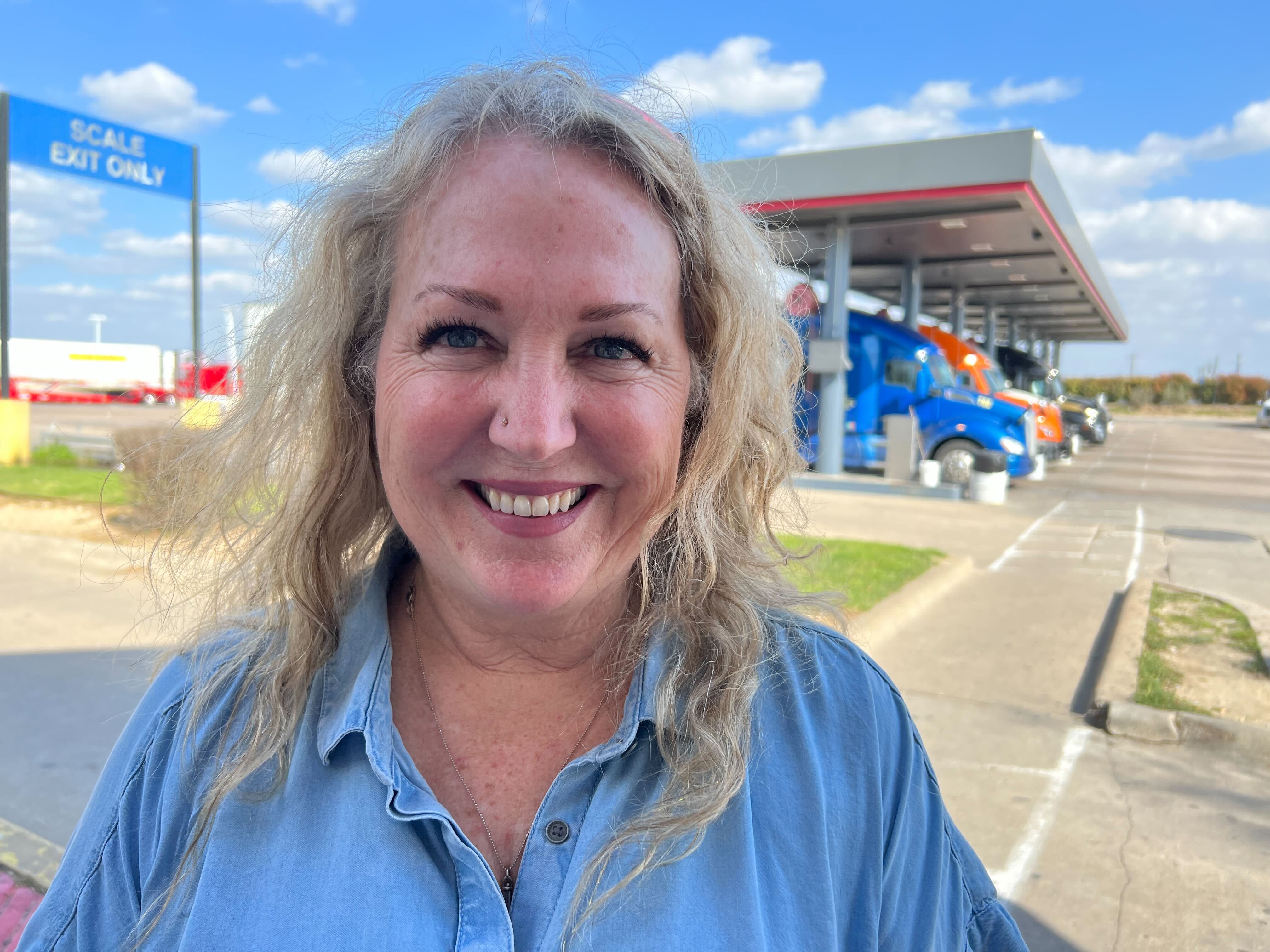 A woman smiles for the camera, a row of big rig trailer unites seen under an awning behind her. She's outside. She's blonde and is wearing a light blue button-up shirt and she sports a nose ring. This is Kellylynn McLaughlin.