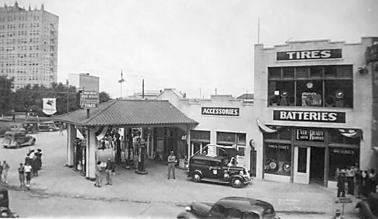 A historical, black and white photo of an old auto service station is seen. A classic, 1950s car is pulling our from under an awning where gas pumps are seen. The signs on the building by it read "Tires, Batteries, and "Accessories." This is the Ever-Ready Auto Service Station in Midland.