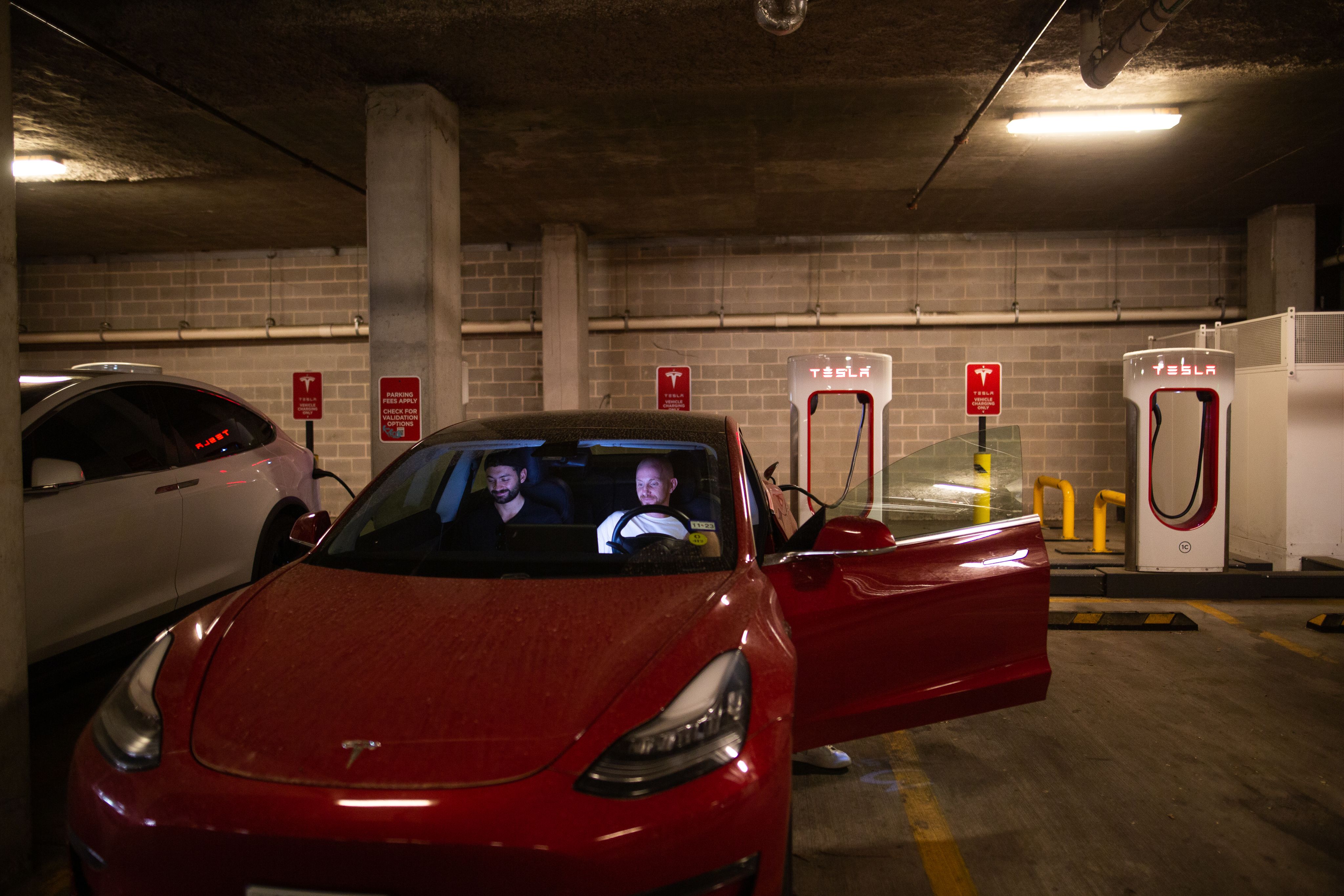 Two men sit in their red Tesla as they wait for the vehicle to charge. They're in a parking garage, the Tesla chargers seen in the back. The driver's side is open and their faces appear to be illuminated by the car interior display or their smartphones.