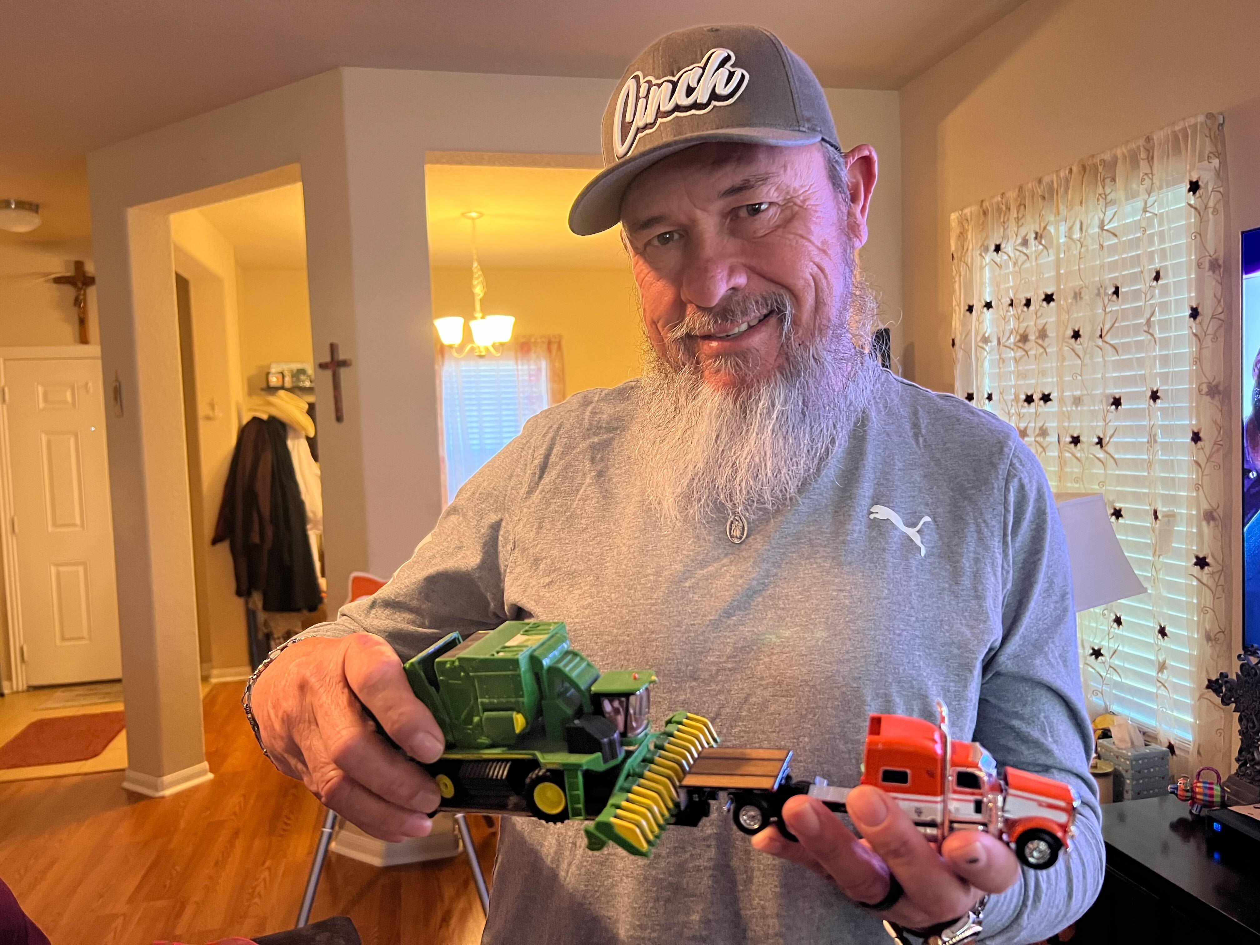 A man holds up a miniature of a big rig truck. The red trailer unit has green farming equipment in tow. He holds up the miniature and smiles at the camera. He's wearing a trucker had, a grey long sleeve shirt, and sports a grey beard. This is truck driver Tony Bergara.