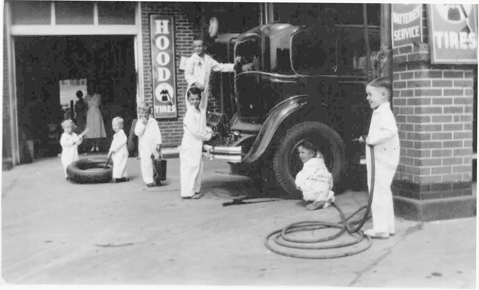 A historical, black and white photo is seen of six children dressed in white posed as if they were working at the Ever-Ready Auto Service Station. They appear to range in age from toddler to about 6-years-old. One child is posed airing up the rear passenger tire of an old, 1950s-era car while another looks under the hood.
